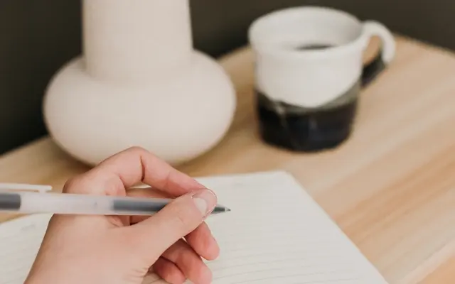 A hand writing in a notebook next to a cup of coffee